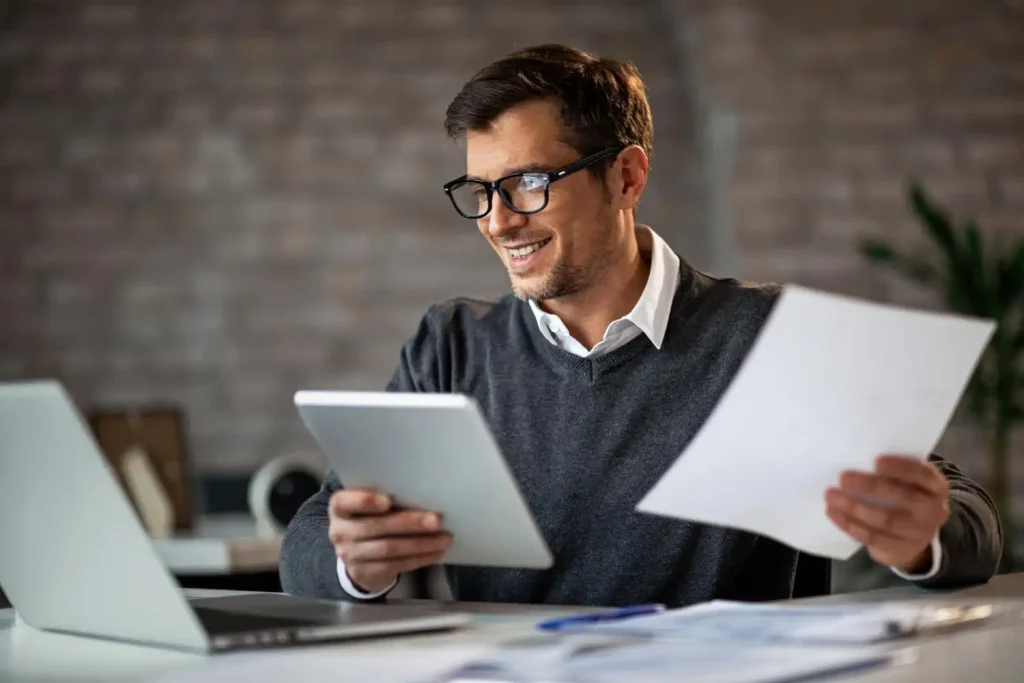 Homem feliz trabalhando de casa usando tablet e computador com foco na produtividade e estratégias de home office para manter o foco remotamente.