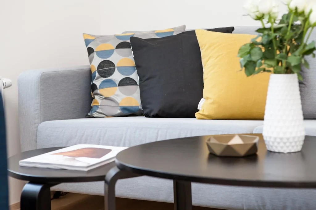 Sala de estar moderna com sofá cinza, almofadas decorativas em cores amarelo, preto e com padrão de círculos, além de uma mesa com revistas, e uma planta em vaso branco ao lado.
