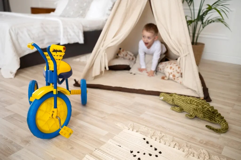 Criança pequena brincando no tapete do quarto infantil organizado e seguro, com tenda de tecido e brinquedos espalhados no chão de madeira clara, ao lado de cama e almofadas
