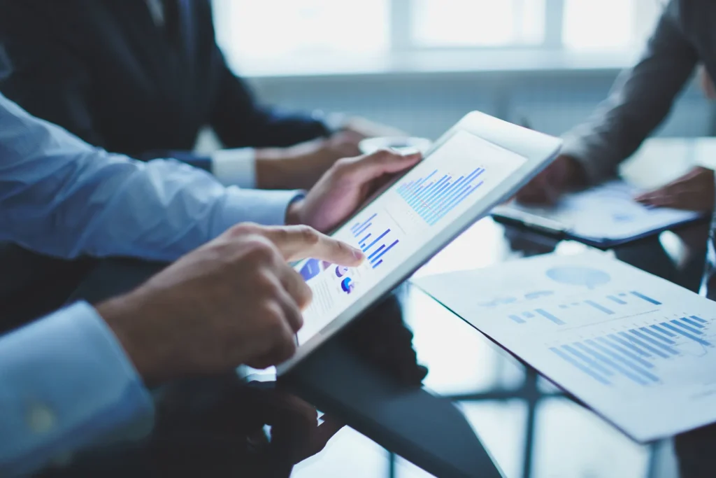 Bastidores da Gestão de Pessoas: reunião entre profissionais em uma mesa, analisando gráficos e relatórios em tablets e documentos, com foco em engajar colaboradores.