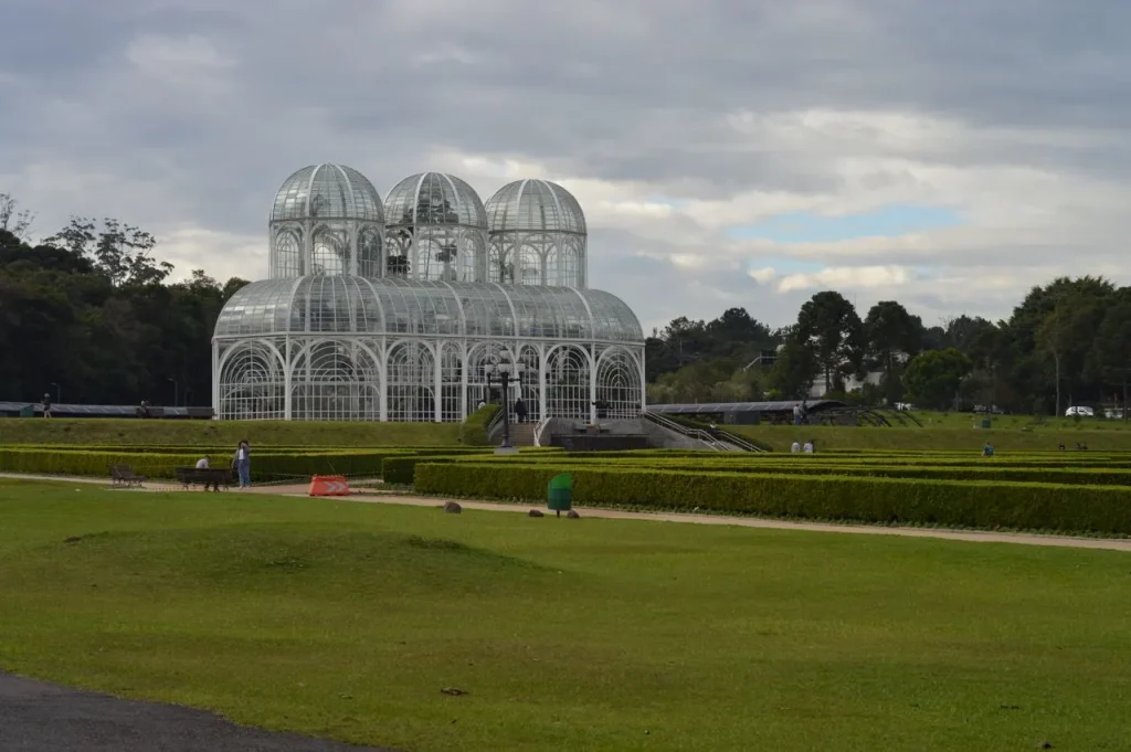 Jardim botânico de Curitiba ao ar livre, com estufa de vidro e cúpulas metálicas ao fundo, cercado por gramados e áreas ajardinadas, em dia nublado.